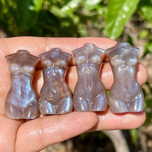 Pode incluir: Quatro pequenas figuras de pedras preciosas esculpidas em forma de torso feminino. As figuras s&atilde;o uma mistura de roxo e cinza com flashes iridescentes azuis e brancos. As figuras s&atilde;o seguradas em uma m&atilde;o contra um fundo verde desfocado.