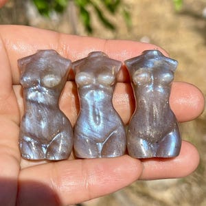 Pode incluir: Tr&ecirc;s pequenas figuras de pedra preciosa esculpidas em forma de torso feminino. As est&aacute;tuas s&atilde;o uma mistura de cinza e azul iridescente, com superf&iacute;cies lisas e polidas. As figuras s&atilde;o seguradas em uma m&atilde;o, com um fundo desfocado.