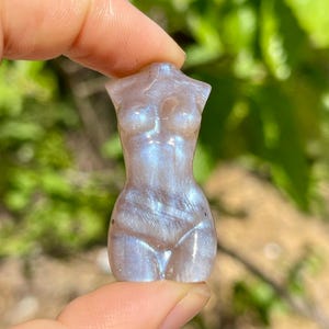 Pode incluir: Uma pequena figura esculpida de um torso humano. A escultura &eacute; de cor cinza claro com tons iridescentes azuis e roxos. A figura &eacute; segurada entre dois dedos contra um fundo verde desfocado.