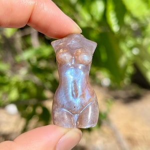 Pode incluir: Pequena estatueta esculpida em pedra preciosa em forma de torso feminino. A pedra tem uma qualidade perolada e iridescente com tons de azul, roxo e castanho. A estatueta &eacute; segurada entre os dedos contra um fundo verde desfocado.