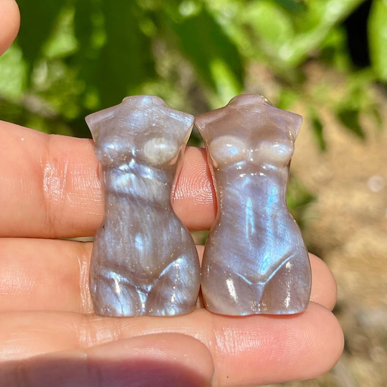 Pode incluir: Duas pequenas esculturas de torso de pedras preciosas iridescentes. As esculturas t&ecirc;m a forma de um corpo feminino, com tra&ccedil;os suaves e arredondados. A cor &eacute; uma mistura de azul p&aacute;lido, cinza e branco, com um efeito brilhante. As esculturas s&atilde;o seguradas em uma m&atilde;o.