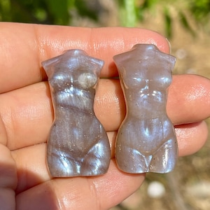 Pode incluir: Duas pequenas figuras de pedra preciosa esculpidas em forma de torso. As figuras s&atilde;o cinza perolado com flashes iridescentes azuis e brancos. As figuras s&atilde;o seguradas na m&atilde;o de uma pessoa.