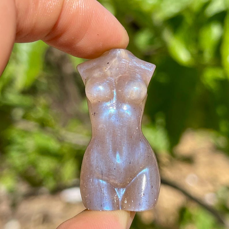 Pode incluir: Pequena estatueta esculpida de um torso feminino. A escultura &eacute; feita de uma pedra cinza clara com flashes iridescentes azuis e roxos. A figura &eacute; segurada entre dois dedos contra um fundo verde desfocado.