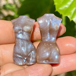Pode incluir: Duas pequenas esculturas iridescentes da forma feminina. As figuras s&atilde;o esculpidas em pedra com brilho perolado, exibindo tons de cinza, azul e castanho. As esculturas s&atilde;o seguradas em uma m&atilde;o, com um fundo verde desfocado. Aproximadamente 5 cm de altura.