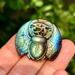 May include: A polished labradorite gemstone carved into the shape of a scarab beetle. The stone displays iridescent flashes of blue, green, and gold, with a detailed relief carving of the insect's body and wings against a circular background.