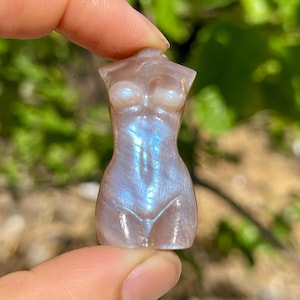 Könnte beinhalten: Kleine, geschnitzte Figur einer weiblichen Form. Die Skulptur hat eine hellbraune Farbe mit irisierenden blauen und weißen Reflexen. Die Figur wird zwischen Fingern vor einem verschwommenen grünen Hintergrund gehalten.