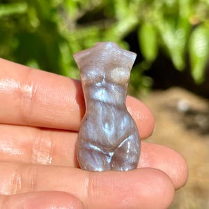 Pode incluir: Uma pequena estatueta esculpida de uma forma feminina. A escultura &eacute; de cor cinza claro com veios iridescentes azuis e brancos. A figura &eacute; segurada em uma m&atilde;o, com um fundo verde desfocado.