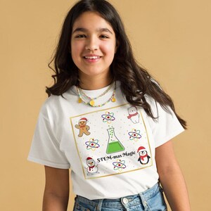 Peut inclure: Un t-shirt blanc avec un motif festif affichant le texte "STEM-mas Magic". Le motif comprend un bonhomme en pain d'épices, un bonhomme de neige, un pingouin et un ours, tous portant des chapeaux de Père Noël, ainsi que des symboles d'atomes et une fiole. Le t-shirt est porté par une personne.