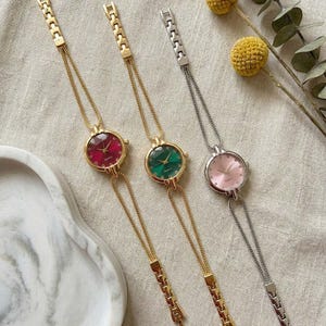 May include: Three pendant watches with round faces in red, green, and pink. The watch faces are set in gold and silver-toned metal. The watches hang from delicate chains with decorative clasps. The background is a neutral fabric.