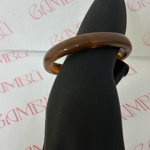 May include: A brown, tortoiseshell-patterned bangle bracelet. The smooth, glossy surface reflects light, showcasing the warm tones of the material. The bracelet is displayed on a black mannequin hand, highlighting its shape and design. Jewelry.