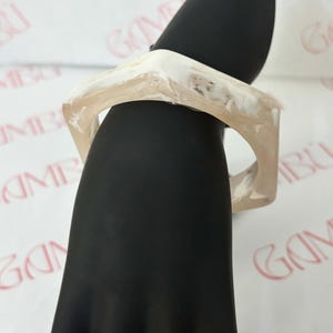 May include: A square-shaped bracelet with a marbled design. The bracelet is a light beige color with white marbling. The bracelet is displayed on a black hand-shaped form against a white background.