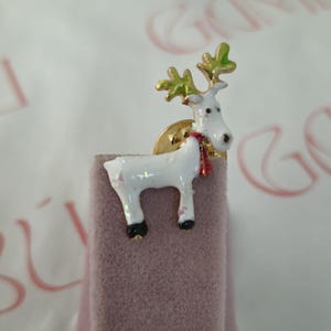 May include: A white reindeer brooch with gold antlers and a red scarf. The reindeer has black hooves and is set on a gold base. The brooch is displayed on a pink velvet surface.