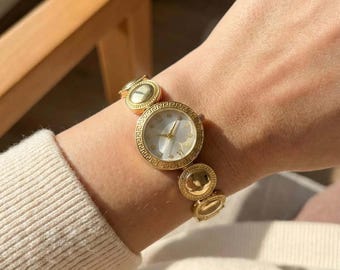 Reloj con pulsera de medallón dorado, esfera con diseño de llave griega y acero inoxidable con circonitas.