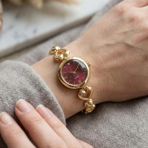 May include: A gold-toned quartz watch with a burgundy face and a heart-shaped link bracelet. The watch has a small diamond accent at the 12 o'clock position. The watch is a stylish accessory.