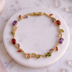 Puede incluir: Pulsera dorada con diseño de vid adornada con gemas de colores. La pulsera presenta gemas de corte ovalado en tonos morado, rojo, naranja, verde y melocotón. El cierre es visible en la parte superior.