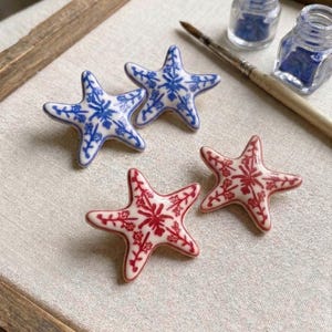 May include: Four ceramic starfish decorations. Two are white with blue floral patterns, and two are white with red floral patterns. The starfish are arranged on a neutral-coloured surface, with paint and brushes in the background.