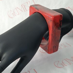 May include: A square red and black marbled resin bracelet. The bracelet is a bold, geometric design, with a glossy finish. The bracelet is shown on a black mannequin hand.