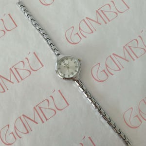 May include: A silver wristwatch with a round face and a linked strap. The watch face has a light-coloured background with black numerals and hands. The strap is made of small, rectangular silver links.