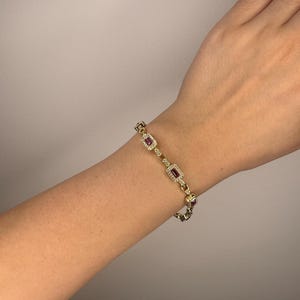 May include: A gold-toned link bracelet featuring rectangular-cut red stones set within frames of small, clear, sparkling stones. The bracelet consists of repeating segments connected by small gold-toned links, creating a continuous chain design.