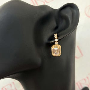 May include: Gold-toned drop earrings with a rectangular, emerald-cut center stone surrounded by smaller, clear stones. The earrings have a post-and-stud closure and are displayed on a black mannequin head. The overall design is elegant and suitable for formal occasions.