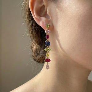 May include: Gold-toned drop earrings featuring a cascade of colorful gemstones. The earrings showcase a variety of shapes and hues, including green, pink, red, blue, and yellow, creating a vibrant and eye-catching accessory.