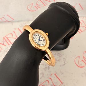 Puede incluir: Un reloj tipo brazalete dorado con caja ovalada, esfera blanca, números romanos negros y manecillas azules. La esfera muestra el texto CHOPPERS y TIMELESS. La correa es un brazalete rígido de metal dorado con acabado pulido.