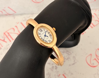 May include: A gold-tone bangle watch featuring an oval-shaped case with a white dial, black Roman numeral hour markers, and blue hands. The watch face displays the text CHOPPERS and TIMELESS. The band is a solid, polished gold-tone metal cuff.