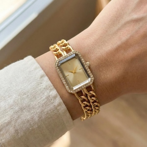 May include: Gold-toned wristwatch with a rectangular face and a chain-link band. The watch face is surrounded by small, sparkling crystals. The band is a double chain design. The watch is worn on a wrist.