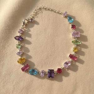 Puede incluir: Una pulsera de cadena en tono plateado con una variedad de piedras preciosas facetadas de múltiples colores y formas, incluyendo tonos morados, rosas, amarillos, verdes y azules. La pulsera incluye un cierre de mosquetón y una cadena extensora.