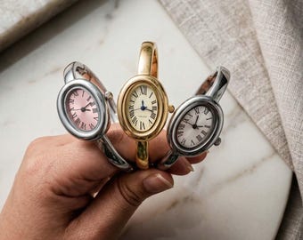 Könnte beinhalten: Drei ovale Armbanduhren in Silber- und Goldtönen. Die Zifferblätter der Uhren zeigen römische Ziffern und Zeiger. Eine Uhr hat ein rosa Zifferblatt, eine ein weißes Zifferblatt und eine ein cremefarbenes Zifferblatt. Die Uhren werden vor einer Marmoroberfläche gehalten.