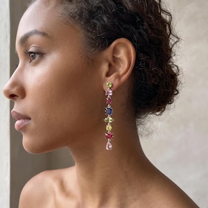 May include: A long drop earring featuring a vertical arrangement of eight multi-colored gemstones in various shapes, including rectangular, oval, and pear-cut stones in shades of lime green, pink, red, and deep blue, set in a silver-toned metal frame.