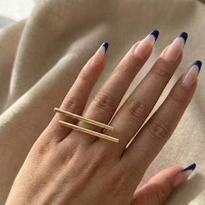 May include: A gold-colored ring with two parallel bars across the finger. The ring is worn on a hand with long nails painted with a French manicure design, featuring a clear base and navy blue tips. The background is a neutral beige fabric.