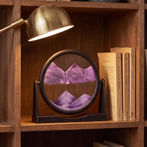 May include: A circular sand art display featuring purple sand layered within a black frame. The frame is mounted on a black rectangular base, allowing it to rotate. The sand forms mountain-like peaks and valleys inside the clear glass enclosure.