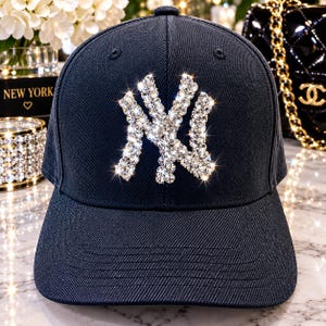May include: A dark navy blue baseball cap featuring a prominent NY logo on the front panel. The logo is constructed from numerous clear, sparkling rhinestones that catch the light. The cap has a structured crown, a curved brim, and stitched eyelets.