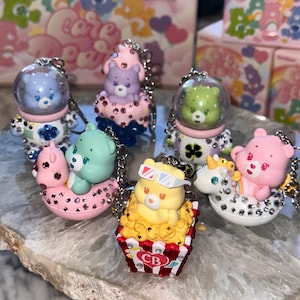 Peut inclure: Une collection de porte-clés sur le thème des Bisounours. Les porte-clés présentent divers Bisounours dans différents décors, notamment des casques spatiaux, du pop-corn et des bouées. Les porte-clés sont ornés de strass et sont attachés à des chaînes argentées.