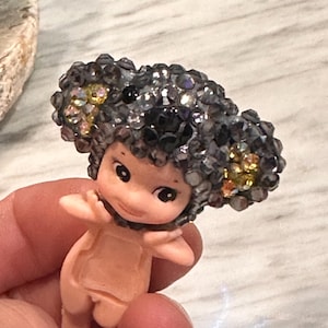 Puede incluir: Una peque&ntilde;a figura de un beb&eacute; desnudo con un tocado enjoyado. El tocado es principalmente gris con acentos dorados y negros. La figura tiene una expresi&oacute;n dulce y se sostiene en una mano.
