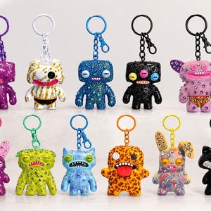 May include: Eleven unique monster-themed keychains featuring various colours, textures, and patterns. Each figure is adorned with small embedded crystals and attached to a metal keyring. The collection includes diverse shapes, expressions, and vibrant finishes.