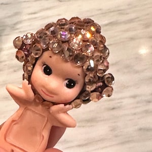 Puede incluir: Una peque&ntilde;a figura de piel clara con grandes ojos negros y una suave sonrisa. El cabello de la figura est&aacute; adornado con numerosos cristales brillantes de color rosa. Los brazos est&aacute;n extendidos y el cuerpo tiene un recorte rectangular.