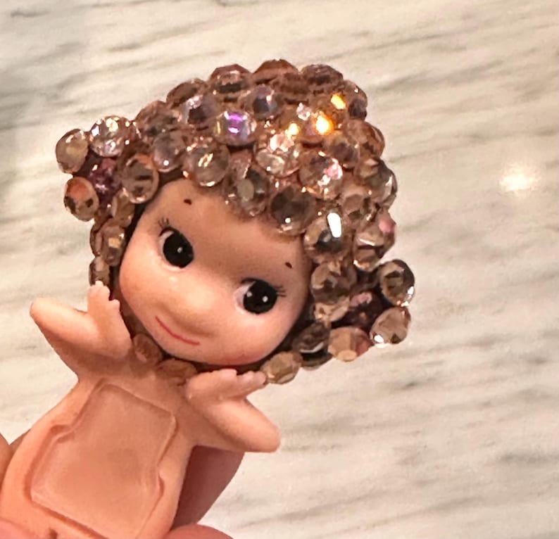 Puede incluir: Una peque&ntilde;a figura de piel clara con grandes ojos negros y una suave sonrisa. El cabello de la figura est&aacute; adornado con numerosos cristales brillantes de color rosa. Los brazos est&aacute;n extendidos y el cuerpo tiene un recorte rectangular.