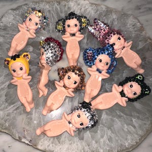 Puede incluir: Una colecci&oacute;n de peque&ntilde;as figuras de beb&eacute;s de color nude con varios peinados de pedrer&iacute;a. Las figuras tienen caras pintadas y est&aacute;n dispuestas sobre una superficie clara y estampada. Algunas tienen sombreros de oso amarillos.