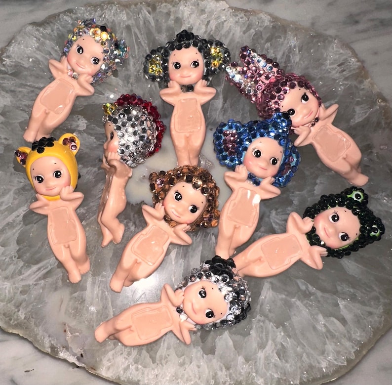 Puede incluir: Una colecci&oacute;n de peque&ntilde;as figuras de beb&eacute;s de color nude con varios peinados de pedrer&iacute;a. Las figuras tienen caras pintadas y est&aacute;n dispuestas sobre una superficie clara y estampada. Algunas tienen sombreros de oso amarillos.