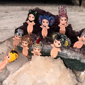 Puede incluir: Una colecci&oacute;n de peque&ntilde;as figuras con forma de mu&ntilde;eca, con cabello brillante y tocados decorativos. Las figuras est&aacute;n dispuestas sobre una geoda con formaciones de cristal morado y blanco. Las mu&ntilde;ecas tienen un tono de piel claro y est&aacute;n en varias poses.