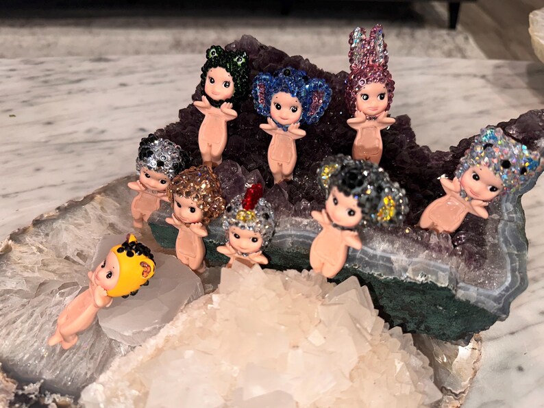 Puede incluir: Una colecci&oacute;n de peque&ntilde;as figuras con forma de mu&ntilde;eca, con cabello brillante y tocados decorativos. Las figuras est&aacute;n dispuestas sobre una geoda con formaciones de cristal morado y blanco. Las mu&ntilde;ecas tienen un tono de piel claro y est&aacute;n en varias poses.