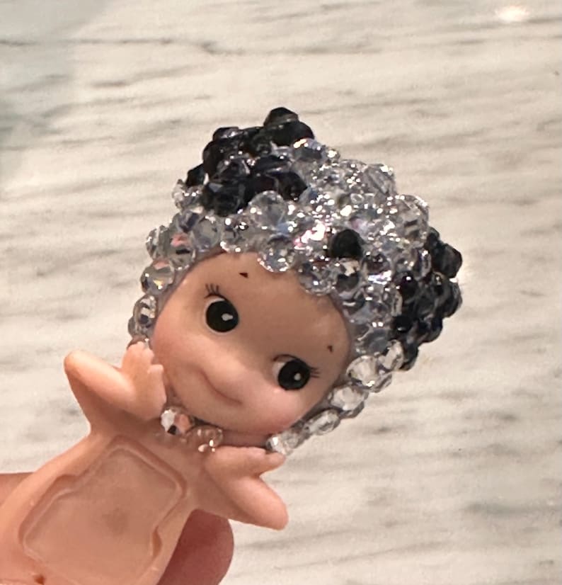Puede incluir: Peque&ntilde;a figura sonriente con tono de piel claro. La figura tiene una cabeza de cuentas negras y transparentes con forma de cristal. La figura tiene ojos negros y brazos extendidos.