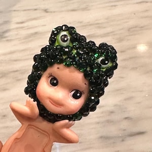 Puede incluir: Una peque&ntilde;a figura de una criatura parecida a una mu&ntilde;eca con un tocado verde con gemas y ojos grandes y expresivos. La figura tiene un tono de piel claro y est&aacute; posada con los brazos extendidos. El tocado se asemeja a la cabeza de una rana.