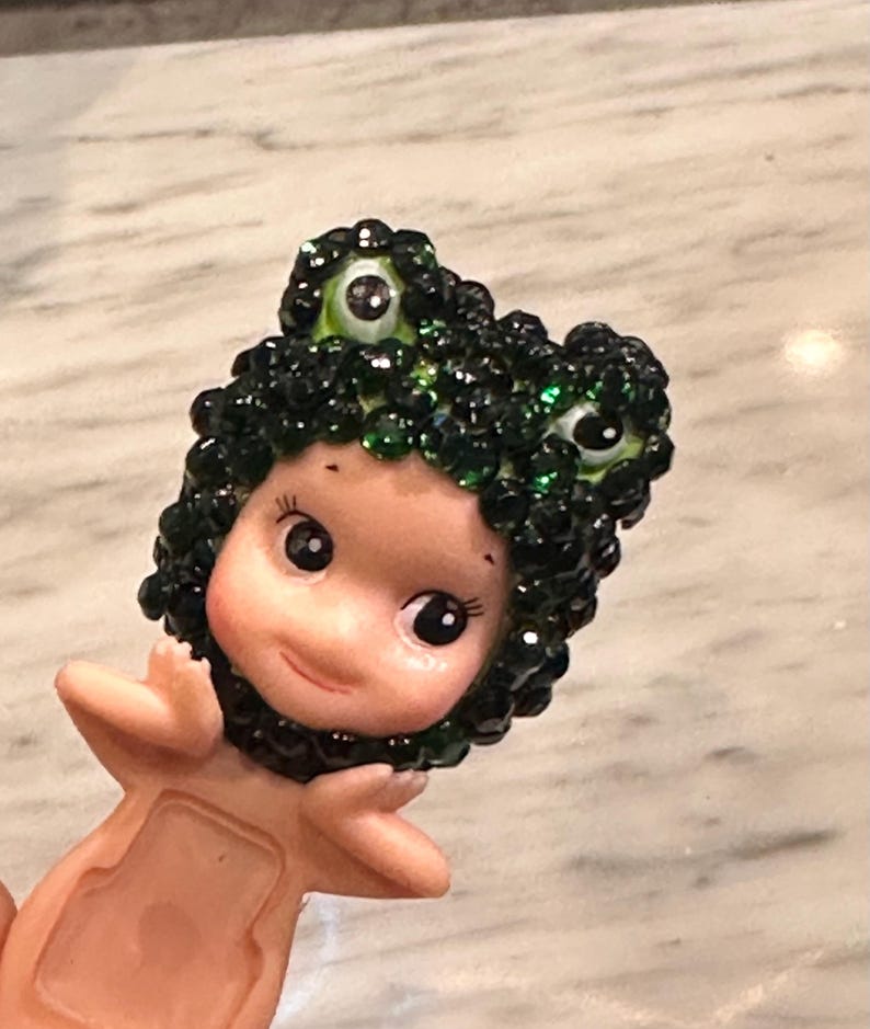 Puede incluir: Una peque&ntilde;a figura de una criatura parecida a una mu&ntilde;eca con un tocado verde con gemas y ojos grandes y expresivos. La figura tiene un tono de piel claro y est&aacute; posada con los brazos extendidos. El tocado se asemeja a la cabeza de una rana.