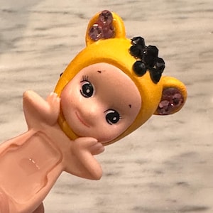 Puede incluir: Peque&ntilde;a figura de color rosa claro de un personaje parecido a un beb&eacute; que lleva una capucha amarilla con detalles en negro y morado. La figura tiene grandes ojos negros y est&aacute; sonriendo. La capucha tiene forma de orejas a los lados.