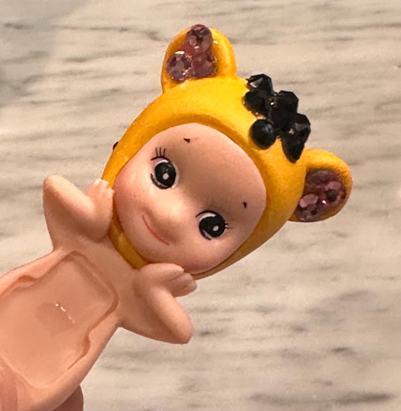 Puede incluir: Peque&ntilde;a figura de color rosa claro de un personaje parecido a un beb&eacute; que lleva una capucha amarilla con detalles en negro y morado. La figura tiene grandes ojos negros y est&aacute; sonriendo. La capucha tiene forma de orejas a los lados.