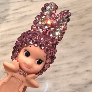 Puede incluir: Figura rosa de un personaje querub&iacute;nico con una diadema enjoyada y cabello rizado. La figura tiene una cara sonriente, ojos grandes y est&aacute; adornada con un collar de joyas. La diadema est&aacute; adornada con gemas iridiscentes.