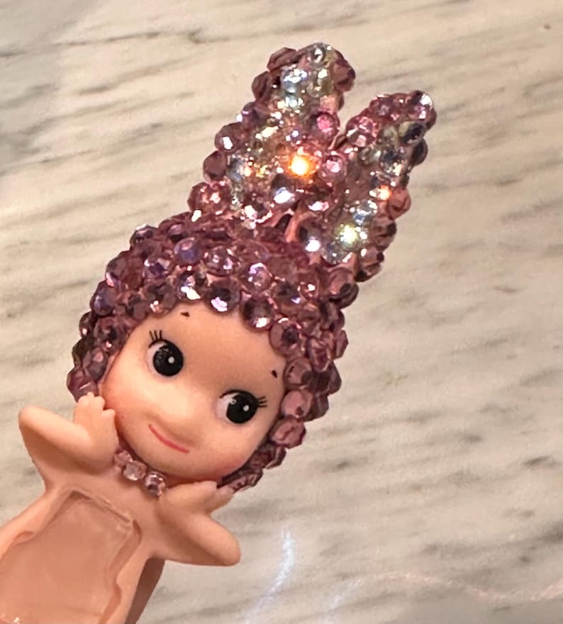 Puede incluir: Figura rosa de un personaje querub&iacute;nico con una diadema enjoyada y cabello rizado. La figura tiene una cara sonriente, ojos grandes y est&aacute; adornada con un collar de joyas. La diadema est&aacute; adornada con gemas iridiscentes.